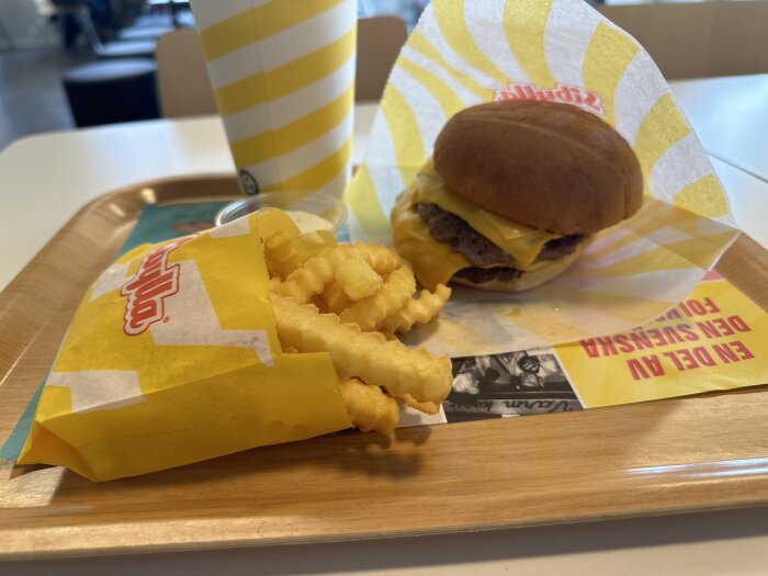 Måltid på Sibylla: en hamburgare med ost, krispiga pommes frites i en gul förpackning och en dryck i en randig bägare, serverad på en bricka.