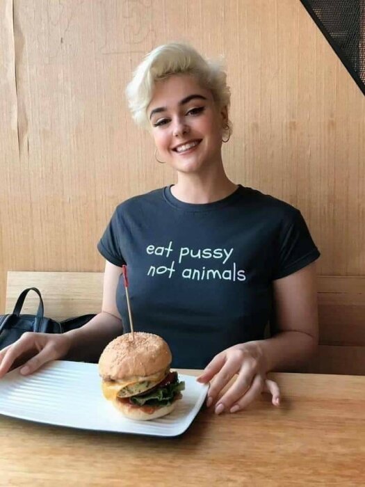 Person med kort blont hår sitter vid ett bord och håller en talrik med en hamburgare. Personen bär en svart tröja med texten "eat pussy not animals".