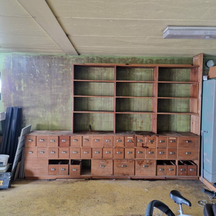 En stor köpmannadisk i trä med flera små lådor och ett hyllsystem ovanpå, placerad mot en grönaktig vägg i ett garage. Några lådor saknas.