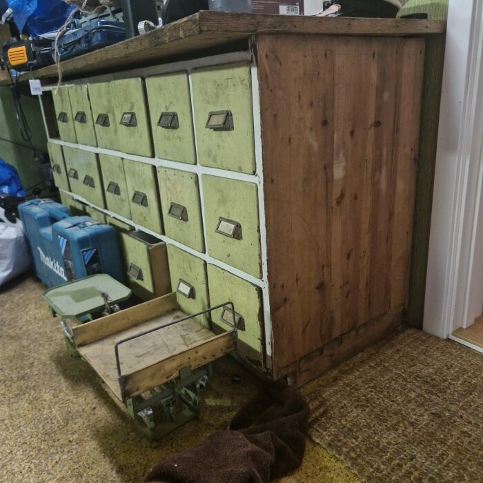 Gammal köpmannadisk i trä med gröna lådor utan handtag, där några av lådorna är utdragna. Disken står i ett garage med diverse verktyg runtomkring.