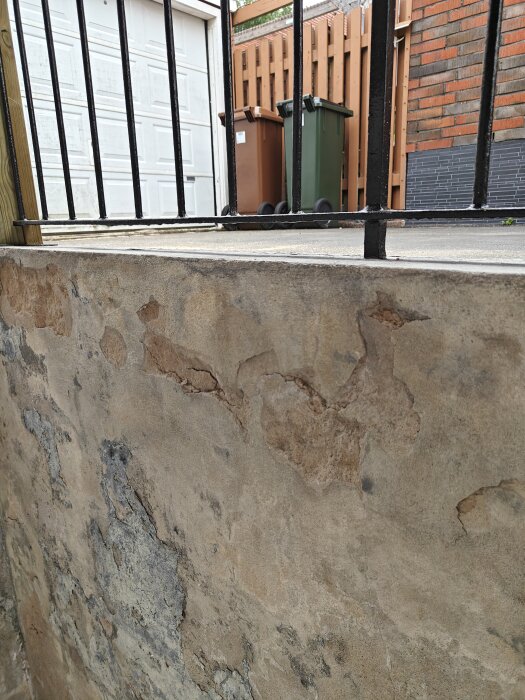 Närbild på en utvändigt renoverad källartrappa med avskavd puts och skador på betongväggen. I bakgrunden syns ett staket, soptunnor och en garageport.