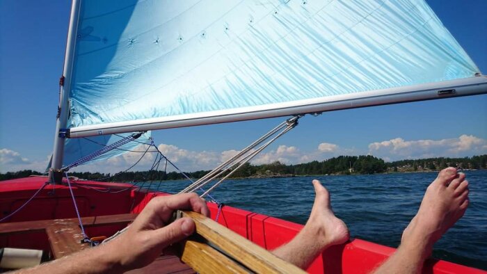 Person seglar en röd båt på en solig dag, hand vid rodret och fötter på båtens kant, med blått segel ovanför och skärgårdslandskap i bakgrunden.