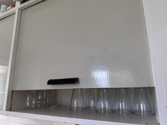 Överskåp i kök från 50-talet med skjutlucka och gamla handtag, flera glas står på hyllan.