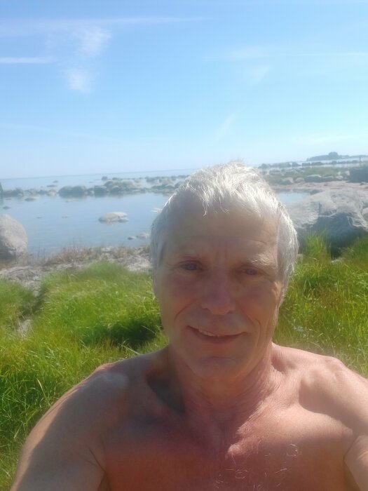 Äldre person utomhus, barbröstad nära grönska vid en stenig strand med havet och en klarblå himmel i bakgrunden.
