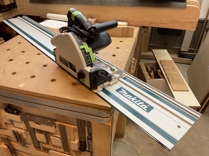 En Festool TS55 kap- och gersåg placerad på en Makita styrskena på ett arbetsbord i en verkstad, omgiven av trästycken och verktyg.