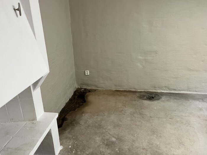 Källarrum med uppbruten parkett, synligt hål i betongplattan och en större blöt fläck i hörnet nära vägg och golv. Väggarna är porösa med misstänkt fukt.