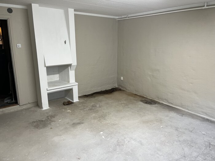 Rum med ljusgrå väggar och ett rivet golv där parkett har tagits bort. Fuktfläckar synliga på golvet. En vit inbyggd hylla finns i hörnet.