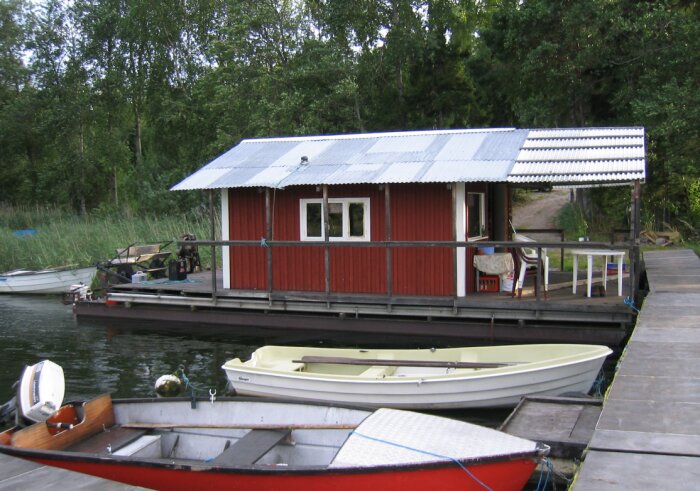 Rött hus byggt på en flotte vid en brygga, omgärdat av båtar och grönska i bakgrunden. Huset har en veranda, och ett metalltak.