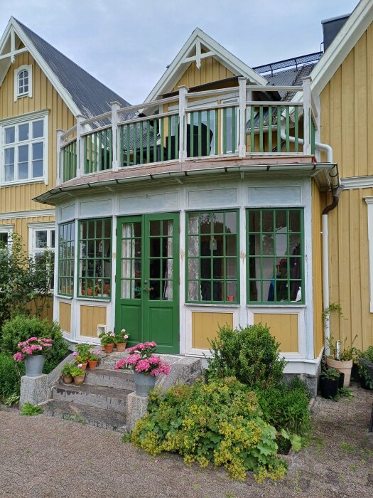 Utvändig tambur med gröna dubbeldörrar och fönster, omgiven av blomkrukor och gröna buskar, på en gulmålad villa med vita knutar och balkong ovanför.