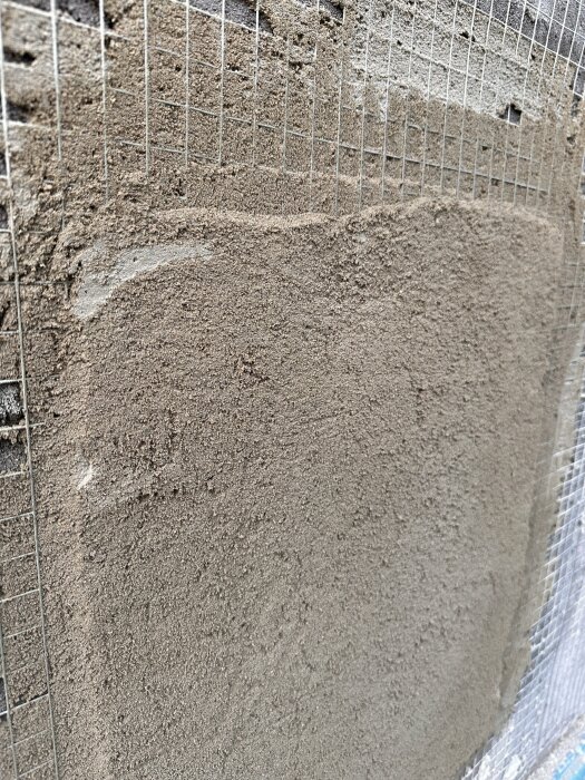 Putsad vägg med nylagd puts som inte är helt torr, synlig armeringsnät bakom puts, oregelbunden yta och några skarpa kanter.