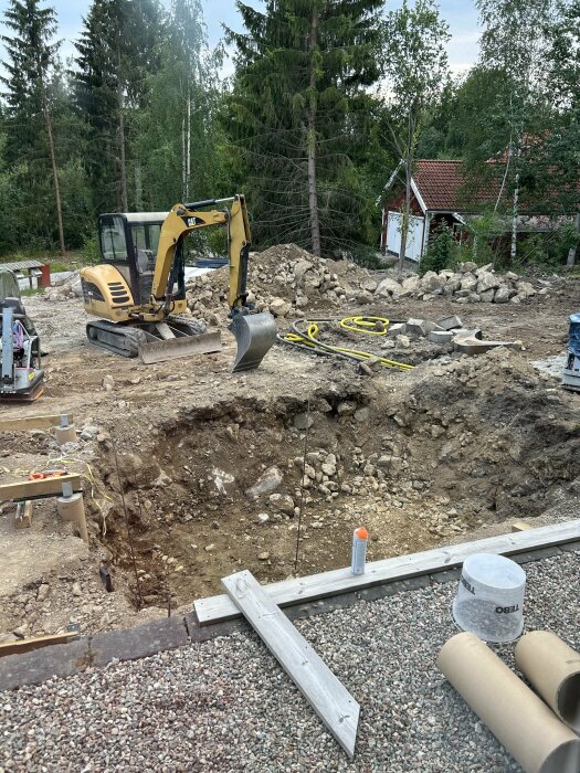 Grävmaskin bredvid stor utgrävd grop på tomt, omgiven av stenar och grus. Verktyg och byggmaterial ligger utspridda i förgrunden. Röd stuga i bakgrunden.