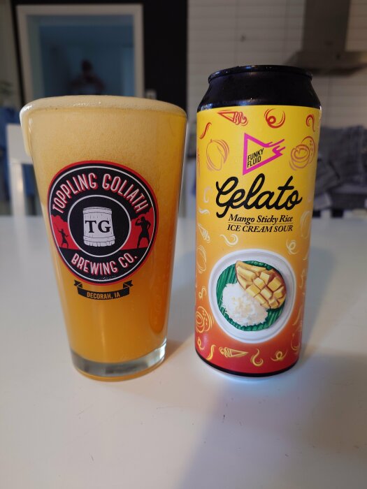 Ett glas fylld med gyllene öl från Toppling Goliath Brewing Co. bredvid en burk Funky Fluid Gelato Mango Sticky Rice Ice Cream Sour.