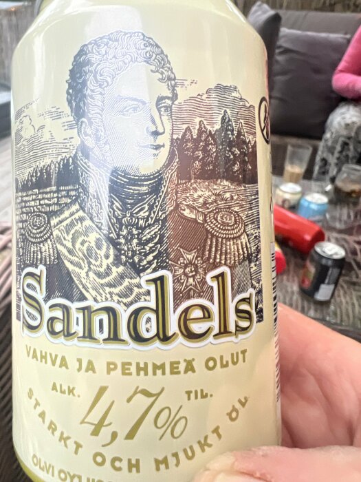 Sandels ölburk med illustration av en man, texten "vahva ja pehmeä olut, alk. 4,7% til., starkt och mjukt öl". Bakgrund med bord, soffor och andra drycker.