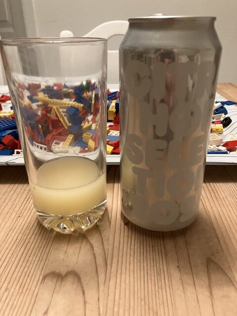 En halvfullt glas med grumlig gul IPA och en ölburk med texten "CITRA HOP SELECTION 003" står på ett träbord, i bakgrunden syns färgglada Legobitar.