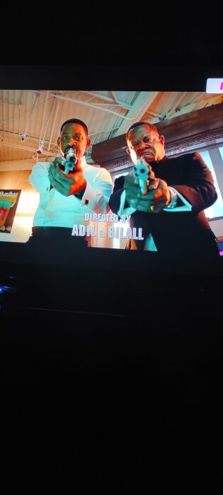 Två män i ljusa skjortor och svarta kostymer riktar pistoler mot kameran. Det står "Directed by Adil & Bilall" på skärmen.