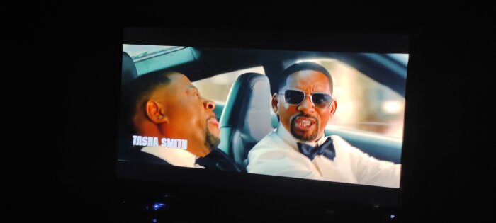 Tv-skärm visar en film med två män i vita skjortor och svarta flugor, sittandes i en bil. Texten "TASHA SMITH" visas i nedre vänstra hörnet.