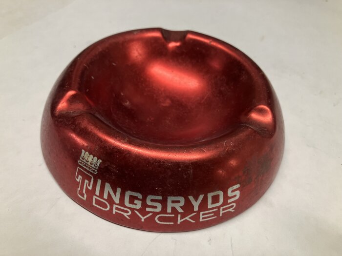 Rött askfat i aluminium med texten "Tingsryds Drycker" och en vit krona, placerat på en vit bakgrund.