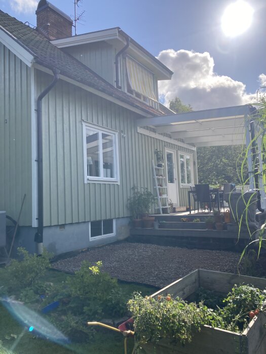 Uteplats under konstruktion nedanför en träveranda. Grusbelagt område förberett för stenmjöl och plattor. Växter och odlingslåda i förgrunden, hus och solsken i bakgrunden.