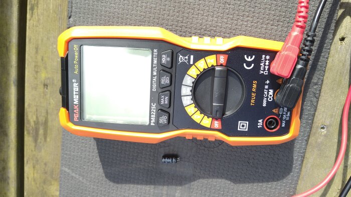 Multimeter PEAKMETER PM8225C med två sladdar anslutna, liggande på en yta bredvid en borttagen kondensator.