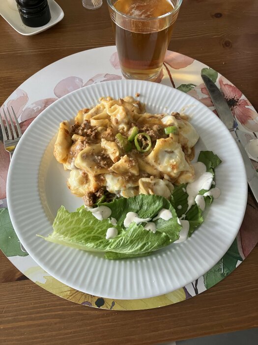 Tallrik med pastagratäng, köttfärssås, skivad sivripaprika och salladsblad med dressing, serverat på en bordstablett. Ett glas dryck står bredvid.