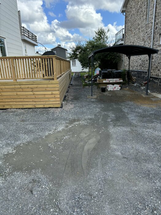 En stenlagd gång med kantsten under konstruktion, skyddad av en altan till vänster och ett stenhustill höger. Byggmaterial och verktyg ses under ett tak.