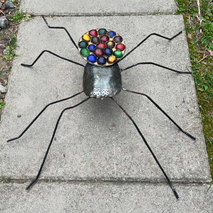 En metallskulptur av en spindel med färgglada glasstenar på ryggen, placerad på en betonggång.