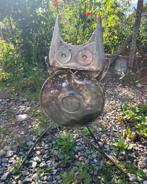 Metallskulpturer av en uggla (en uggla gjord av en bilkåpa med ordet "Saab" och två ögon) och en spindel står på en grusig mark omgiven av växter.