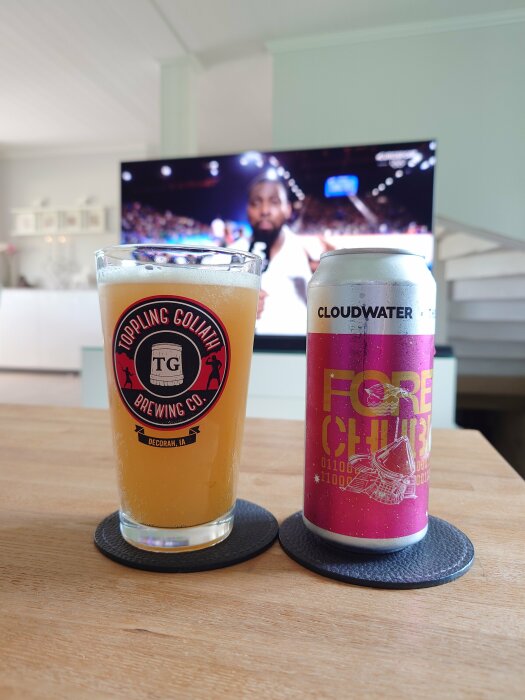 Ett glas med en öl från Toppling Goliath Brewing bredvid en burk öl från Cloudwater x The Veil, stående på ett bord framför en pågående TV-sändning av en judotävling.