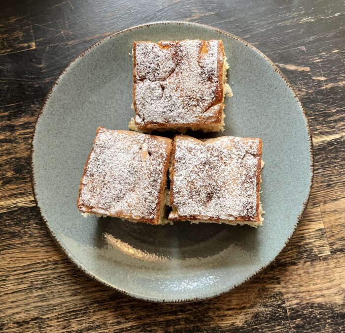 Tre bitar päronkaka med kardemumma och vanilj, pudrade med florsocker, på en grå tallrik placerad på ett bord av trä.