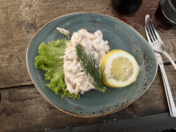 Brödskiva med Skagenröra, garnityr av salladsblad, citronskiva och dill på en blå tallrik, liggande på ett rustikt träbord med bestick och dryck i bakgrunden.