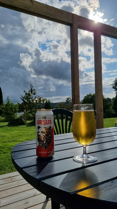 En burk Ursus-öl och ett glas öl på ett bord utomhus, med bakgrund av en halvt molnig himmel och grönska.