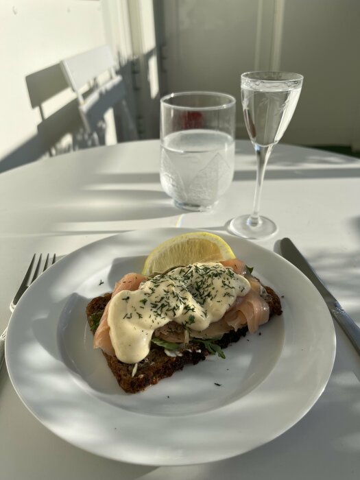 Hemlagat smörrebröd med kallrökt lax, senapsmajonnäs, ruccola och citron på rågbröd. Ett glas vatten och en snaps vid sidan av på ett vitt bord.