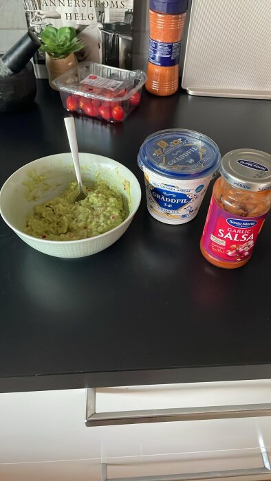 Skål med hemmagjord guacamole, bredvid en burk gräddfil och en burk Santa Maria salsa, på en köksbänk; tomater och kryddor i bakgrunden.