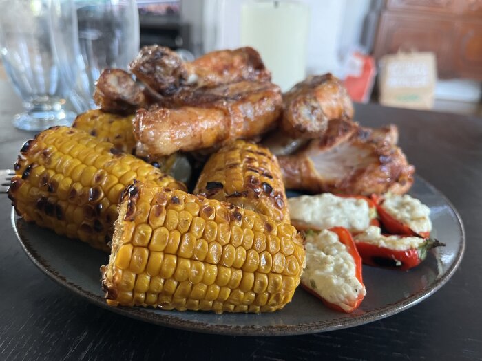 Grillad kyckling, majskolvar, fyllda snackpaprikor med chili på en tallrik vid matbordet; glas i bakgrunden.