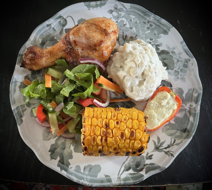 En tallrik med grillad kycklingklubba, majskolv, sallad, potatissallad och en fylld paprika.