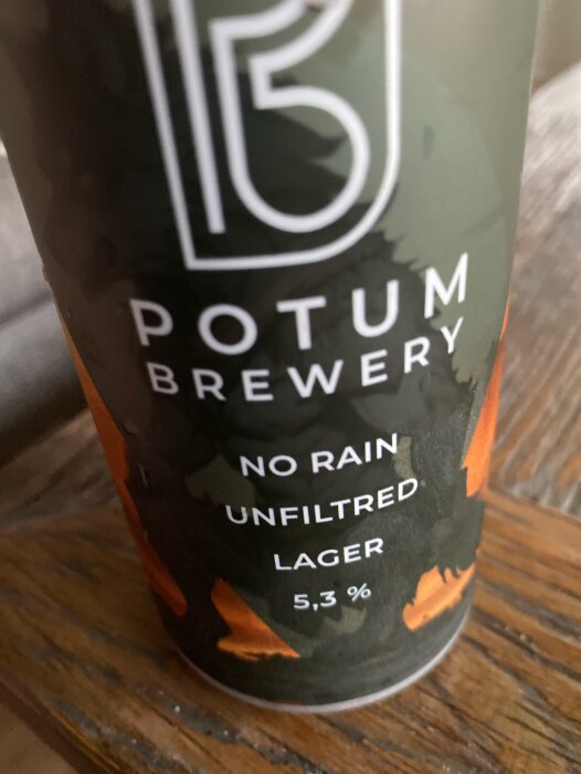 Närbild på en ölburk från Potum Brewery med en etikett som säger "No Rain Unfiltered Lager 5,3%" mot en träbakgrund.