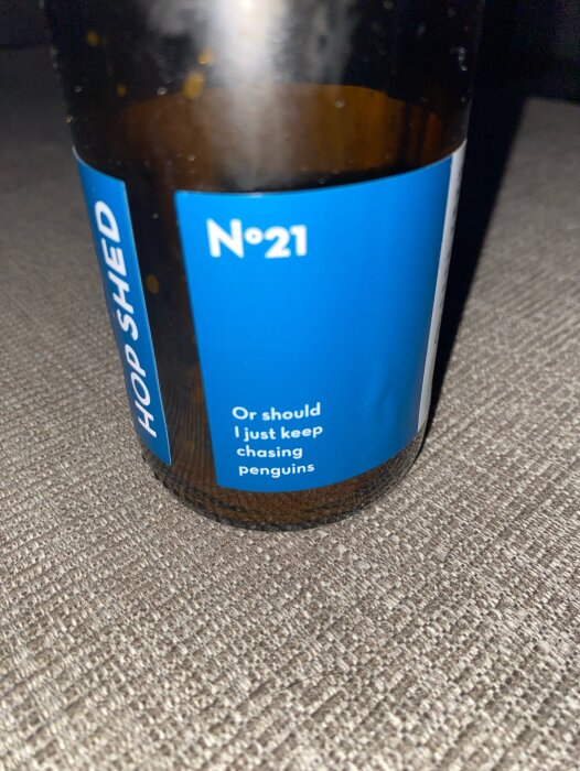 Ölflaska från Hop Shed med blå etikett där det står "No 21. Or should I just keep chasing penguins." Flaskan står på en grå yta.