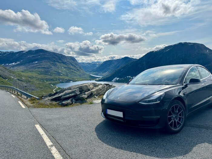 En svart Tesla parkerad vid väg med naturskön utsikt över berg, dalar och en sjö under molnig himmel.