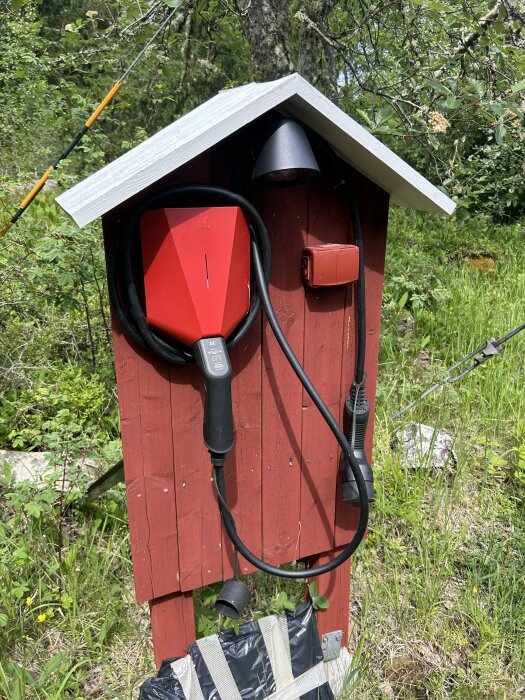 Röd fristående laddbox med tak, utrustad med en svart kabel och en lampa ovanpå, installerad i en grönskande trädgårdsmiljö.