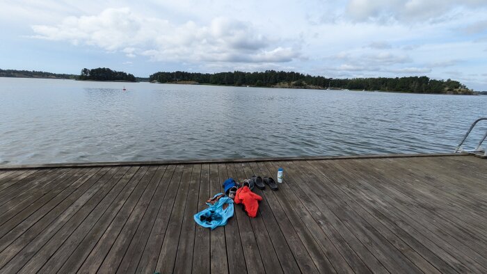 Kläder, skor och en vattenflaska ligger på en brygga vid en sjö med skogsklädda öar i bakgrunden under en molnig himmel.