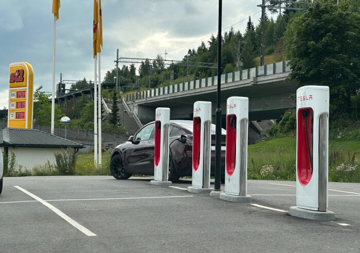 Tom parkeringsplats med Tesla-laddare i Örnsköldsvik. En svart bil är parkerad bredvid laddstolparna. Skylt visar bränslepriser från St1. Bakgrund av bro och träd.