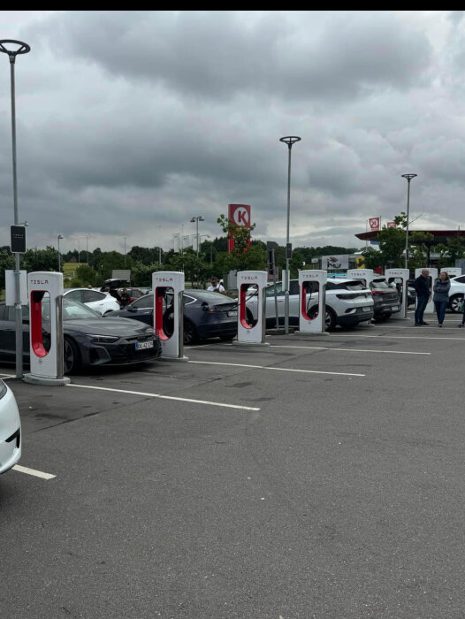 Bilar parkerade vid Tesla-laddstationer på en parkeringsplats, vissa bilar står över flera rutor, ett bensinstation-skylt med "K" syns i bakgrunden.