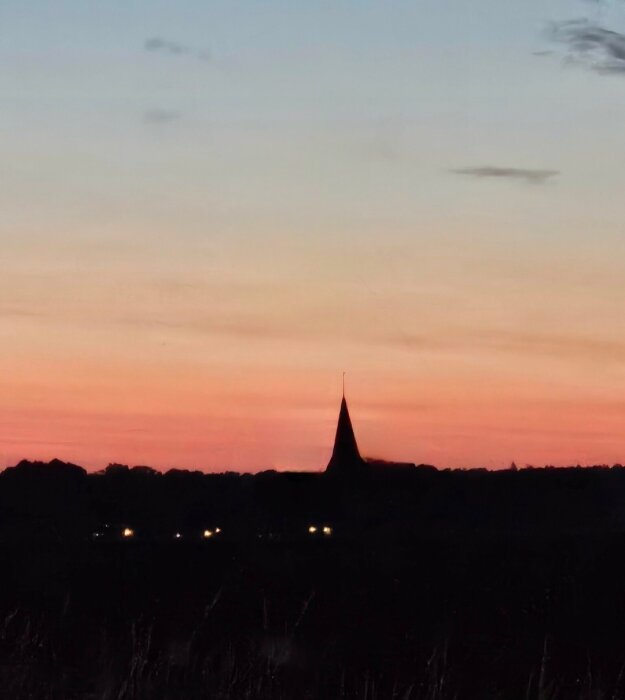 Silhuett av en byggnad med spetsigt tak i solnedgången, upplysta fönster nedtill och en mast på toppen som syns mot himlen.