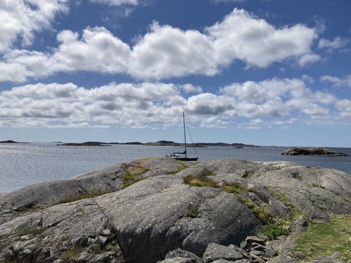 En segelbåt ligger förankrad vid klippor längs en kustlinje under en blå himmel med vita moln.