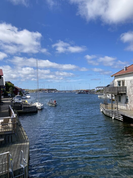 Småbåtar ligger förtöjda längs bryggor i en skyddad vik, med rymliga fastigheter på varje sida och ett öppet hav synligt under en klarblå himmel med lätta moln.