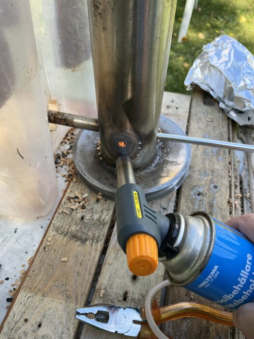 Hand värmer en rökare med en gasbrännare, rökspån och tång synliga på ett träbord utomhus.