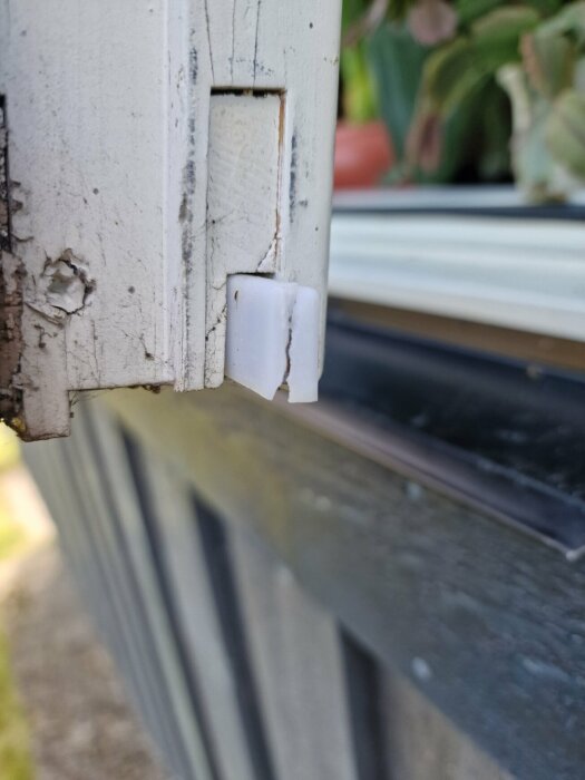 Närbild på en vit plasthylsa i en metallisk fönsterspanjolett där plasthylsan ser sprucken ut. Bakgrunden visar en del av ett hus och några växter.