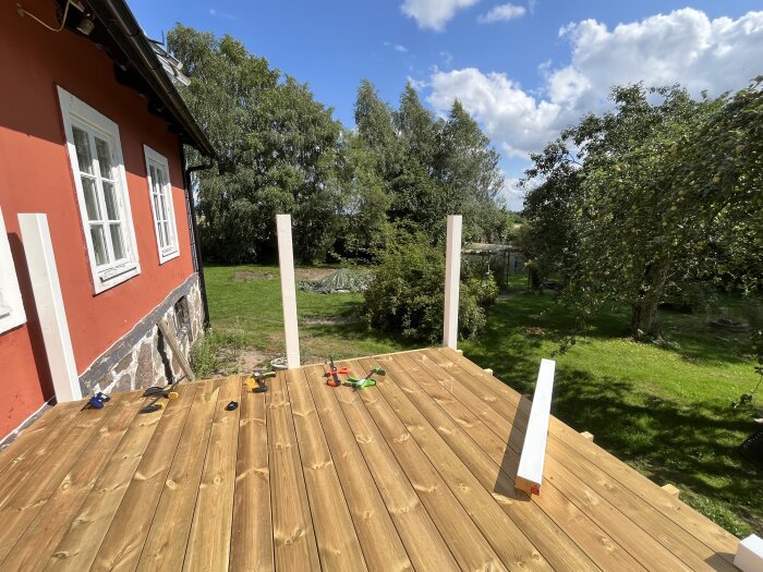 Nybyggd balkong med träplankor, vita stolpar och verktyg på golvet, intill en röd fasad med vita fönster. Grönskande trädgård och blå himmel i bakgrunden.