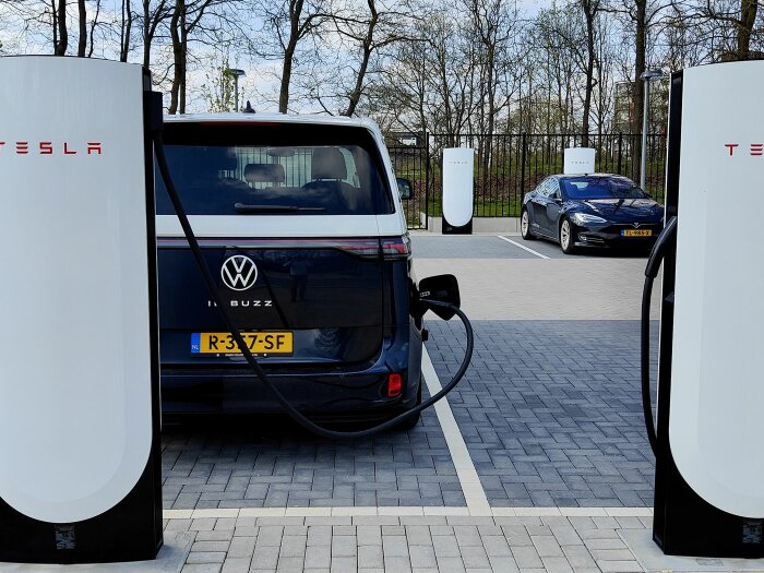 En elbil av märket Volkswagen laddar vid en Tesla-laddstation, med en annan elbil parkerad i bakgrunden.