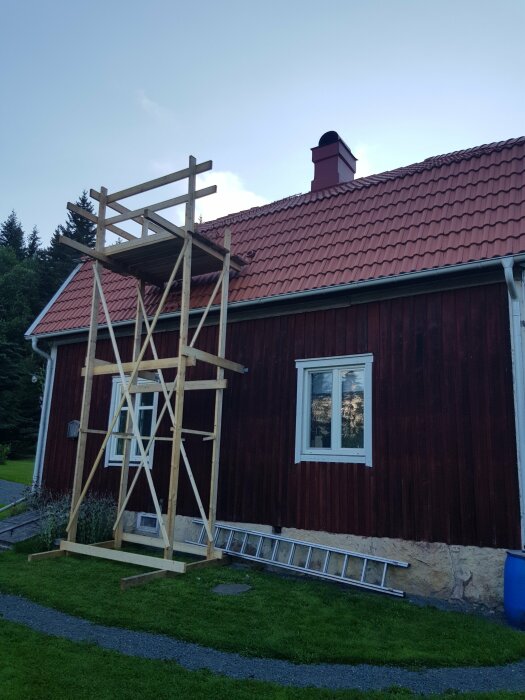 En enklare byggnadsställning av trä står lutad mot ett rött trähus med tegeltak. En stege och en blå tunna står vid sidan av huset.